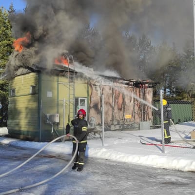 Palomies suihkuttaa vettä pieneen puurakenteiseen taloon, jonka katosta näkyy liekkejä ja yllä on savua. Etualalla toinen palomies kantaa lentkua.