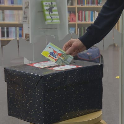 En hand sätter in ett julkort i en svart låda för att senare delas ut till ensamma äldre. I bakgrunden syns biblioteksböckr på hyllor.