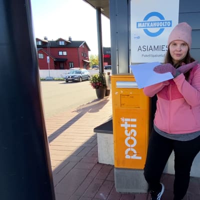 Nainen seisoo postilaatikon edessä kirjeen kanssa kädet ristissä.