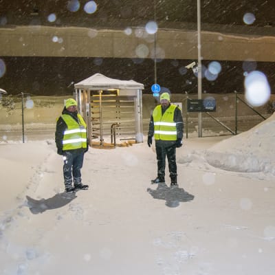 Kaksi henkilöä seisoo lumisen portin edustalla heijastinliivit päällä. 
