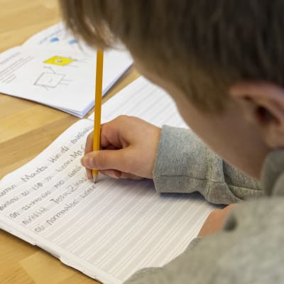 Poika koulussa äidinkielentunnilla kirjoittaa lyijykynällä tarinaa vihkoon tekstaten.