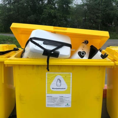 Rivissä kolme keltaista kierrätysmuovin keräysastiaa, joista keskimmäiseen on virheellisesti tungettu valkoinen muovinen matkalaukku.