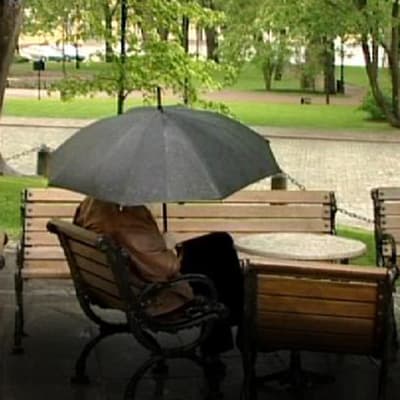 Mies isuu puiston penkillä mustan sateenvarjon alla