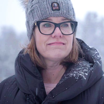 Närbild av Maria Leppäkari ute i snön. Hon har en grå mössa och en svart jacka med snöflingor på kragen. I bakgrunden en snöig skog.