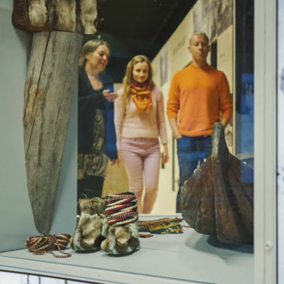 Vitrin med skoltsamernas föremål: liten träbåt, rep, och traditionella pälsskor. I bakgrunden tre personer som tittar på sakerna.