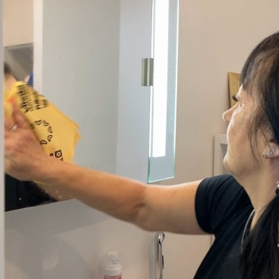 Siivooja Kaija Peni pyhkii kylpyhuoneen peilikaapin ovea keltaisella mikrokuituliinalla.