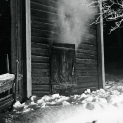 En svart-vit bild på en gammal bastu. Det är vinter och ångan stiger från öppningen till bastun.