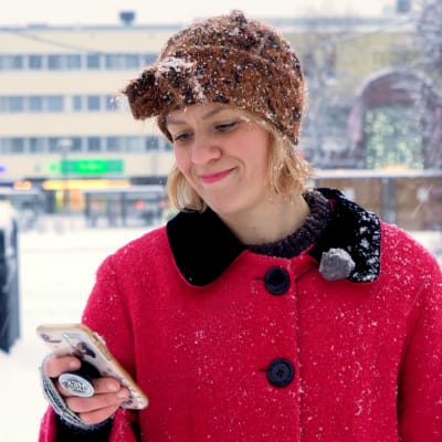En kvinna i röd jacka står på Borgå torg. Det snöar. Kvinnan håller i en mobiltelefon, ler och tittar ner på telefonen.