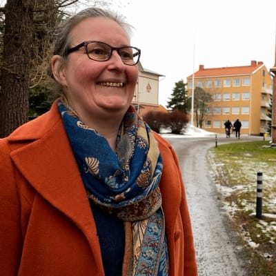 Yliopistotutkija Teija Koskela Rauman Opettajankoulutuslaitoksen pihalla. Taustalla kampuksen rakennukset. Teija hymyille, hänellä on ruosteenoranssi villakangastakkia sekä sinisävyinen kaulahuivi. Hän hymyilee ohi kameran.