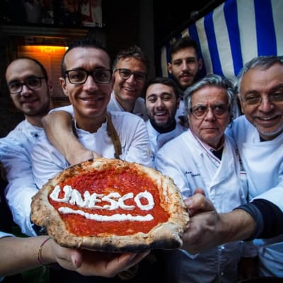 Åtta pizzabagare håller upp en napolitansk pizza där det står skrivet Unesco.