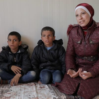 En mamma i röd slöja sitter på en säng i ett flyktingläger tillsammans med sina två små söner 