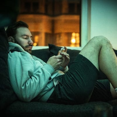 En man ligger på soffa och tittar på sin telefon. 