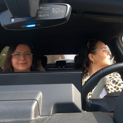 Två kvinnor sitter i en bil. Den ena tittar åt sidan, den andra tittar in i kameran.