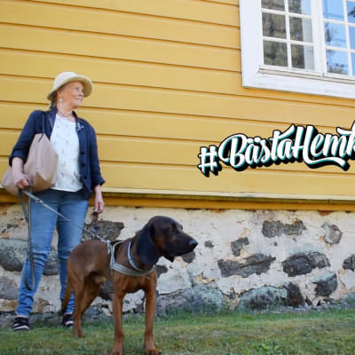 Hedda Kainz står lutad mot en gul gammal trävägg med hunden Buster. På väggen står det bästahemknuten.