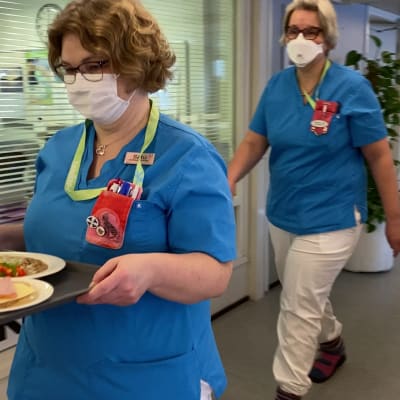 Kaksi siniasuista hoitajaa käytävällä vie ruoka-annoksia potilaille 