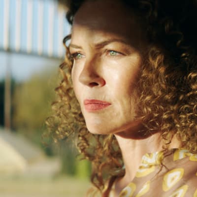 Karaktären Katrina står på en veranda med solen i ansiktet.