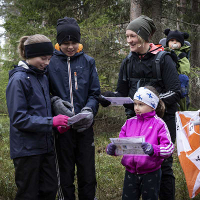 Helmi, Mikael, Heidi, Milo ja Minea Vanhala tutkivat karttojaan metsässä. Oikealla alakulmassa on rastilippu. 