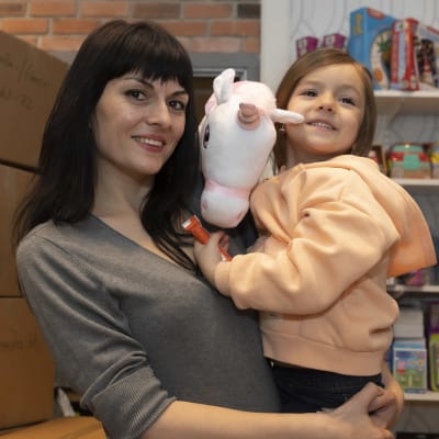 Tummahiuksinen nainen (Ljudmila Dianchenko) ja tyttärensä Viktoria, jolla sylissä yksisarvis-keppihevonen.