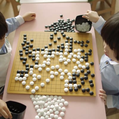 Två barn sitter och spelar go. Det kvadratiska brädet mellan dem är fullt med vita och svarta spelknappar.