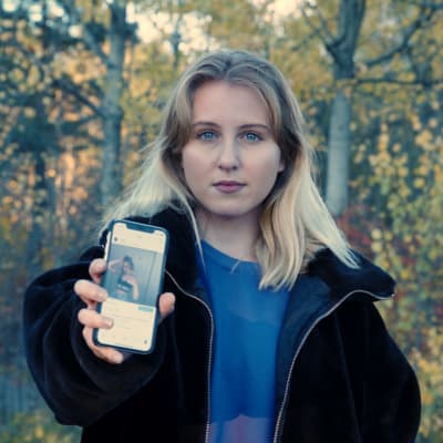 Vaaleahiuksinen nainen seisoo koivumetsän edessä ja näyttää puhelimen ruutua kameralle. Instagram on auki ruudulla.