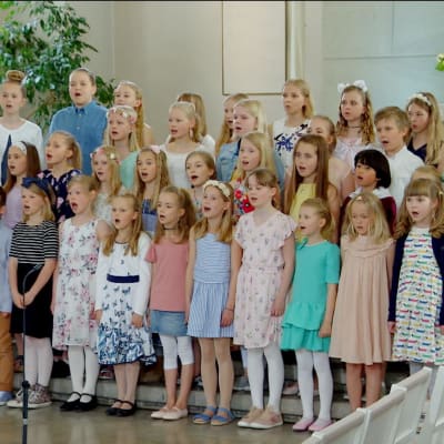 Pastellklädda barn med blommor i håret sjunger framme i en kyrka.