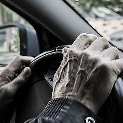 Symbolikuva ikäihmisistä liikenteessä eli vanha mies puristaa auton ohjauspyörää eli rattia käsillään.