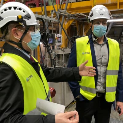 Suojavarusteisiin pukeutunut GTK:n pääjohtaja Kimmo Tiilikainen puhuu Jouko Niemiselle ja heilauttaa samalla kättää Outokummun koetehtaalla.