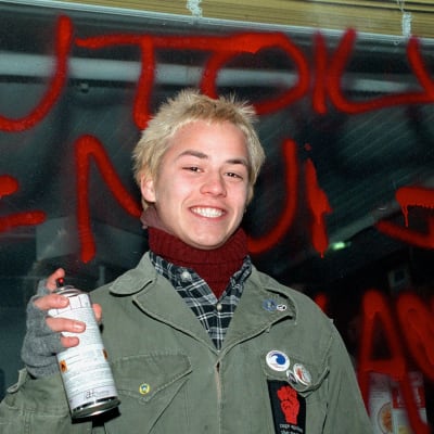 Näyttelijä Petteri Jamalainen seisoo Kotikadun bensiksen edessä kädessään spray-maalipurkki ja takanaan seinäkirjoitus