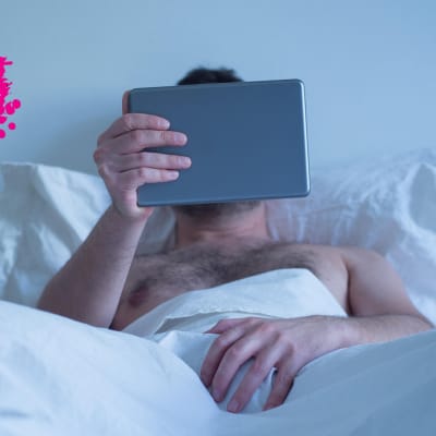 en man son rör sin penis med sin hand på täcket och ser på sin smart tablet