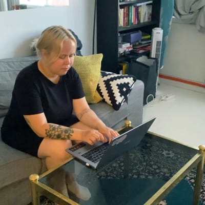 En kvinna med ljust hår och svart tröja sitter i en soffa och jobbar på sin laptop.