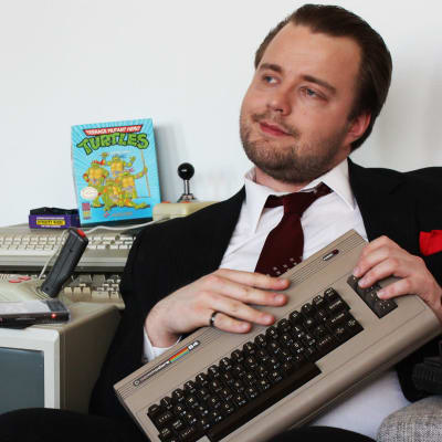 Toimittaja Antti Melkko sliipattuna, pukuun pukeutuneena, vanhan Commodore 64 konsolin kanssa.