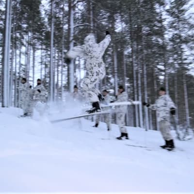 Lumipukuinen kadetti hyppää 360-hypyn metsäsuksilla hyppyristä.
