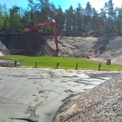 En grävmaskin jobbar med sandvallar som omger en gräsplätt och ett gjutet område.