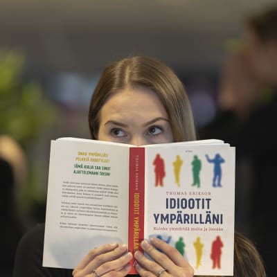 Nainen katsoo kameraan Idiootit ympärilläni -kirjan takaa kyllästynyt ilme kasvoillaan. Takana on himmeällä kaksi muuta hahmoa.