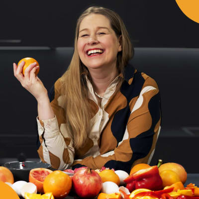 Kirjailija Leena Parkkinen nauraa appelsiini kädessään ja röykkiö vihanneksia ja hedelmiä edessään pöydällä.