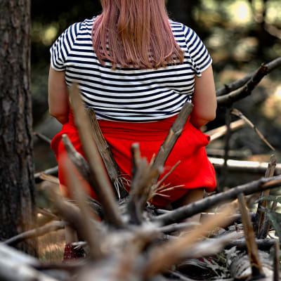 Nainen istuu kaatuneen puun päällä selkä kameraan päin. Puusta törröttää teräviä oksia.