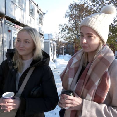 Carita Helminen ja Anniina Halonen seisovat ulkona ja pitelevät kahvikuppeja. Maisema on talvinen ja vasemmalla puolella on työmaakontteja.