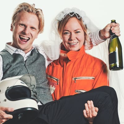 Joppe Dikert iklädd formulaoverall med sin man Oskar Pöysti i famnen. Han har en formel1- hjälm i famnen och en champagneflaska i handen. De ser ivriga och glada ut. 