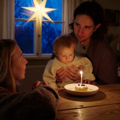 En man, en kvinna och ett litet barn som är i mannens famn. De sitter vid ett köksbord. På bordet finns en tallrik med en minitårta med ett ljus i. I bakgrunden hänger en julstjärna i fönstret.