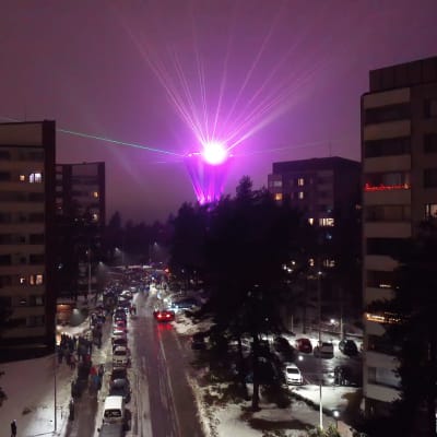 Pimeällä otettu kuva, jonka keskellä menee katu. Kadun varrella on autoja ja runsaasti ihmisiä. Vasemmalla ja oikealla kuvassa on kerrostaloja. Horisontissa näkyy vesitornia, joka hehkuu vaaleanpunaisena ja siitä lähtee vaaleanpunainen valonsäde.