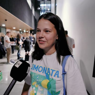 En ung kvinna blir intervjuad med mikrofon. Personen har axellångt mörkt hår och är iklädd en vit t-skjorta med italienskt tryck med texten "Limonata". Personen bär också på en ryggsäck. I bakgrunden syns många personer.