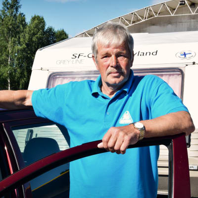 Olof Bussman i klädd en turkos pikéskjorta står lutad mot en bildörr. I bakgrunden skymtar en husvagn.
