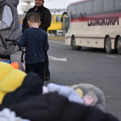 Liten pojke och några vuxna ute på en parkeringsplats vid Ukrainska gränsen i Polen. En låda med leksaker i förgrunden och en buss i bakgrunden.