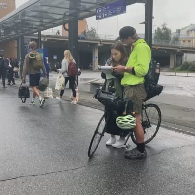 Nainen ja mies seisovat asemalaiturilla. Nainen kirjoittaa lehtiöön, ja mies seisoo vierellä polkupyöränsä kanssa.