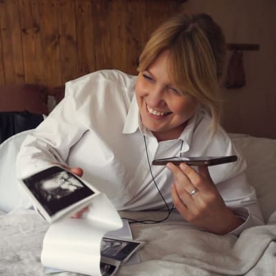 Gravid leende kvinna ligger i sängen och håller i en ultraljudsbild och pratar i telefon.