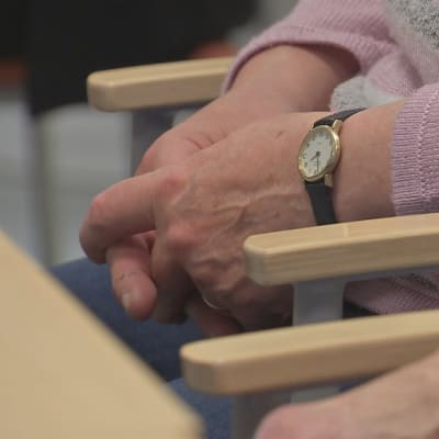 En person med rosa tröja och armbandsur sitter med knäppta händer vid ett bord.
