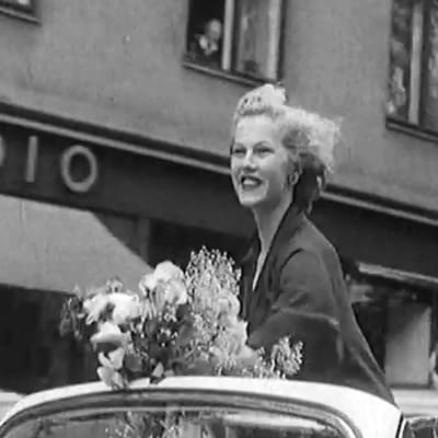 Armi Kuusela vilkuttaa autosta kukkakimppu kädessään. Yleisö on kerääntynyt Helsingin kaduille juhlistamaan Miss Universumia, Armi Kuuselaa.