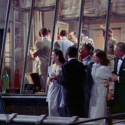 Juhlivia ihmisiä kerrostaloasunnon parvekkeella ja lasin takana sisällä. Kuva Alfred Hitchcockin elokuvasta Takaikkuna.