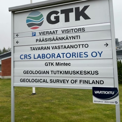 GTK Mintecin tutukimuskeskus ulkoa kuvattuna, etualalla opastekyltti, taustalla rakennus ja pihan poikki kävelevä mies,