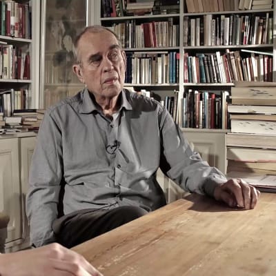 Christoffer Strandberg utklädd till Jörn Donner. Han är sminkad för att se gammal ut samt har stora ögonbryn. Han har också på sig en säckig kavaj. Han sitter och pratar med Jörn i Jörns arbetsrum. I bakgrunden syns bokhyllor fyllda med böcker. 